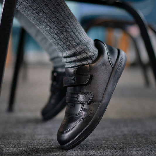 A child in cozy gray socks and Bobux Kid+ LeapTrainer black leather school shoes with sturdy soles and easy fastenings sits on gray carpet. ksnz, 9420068510494, 9420068510500, 9420068510517, 9420068510524, 9420068510531, 9420068510548, 9420068510555