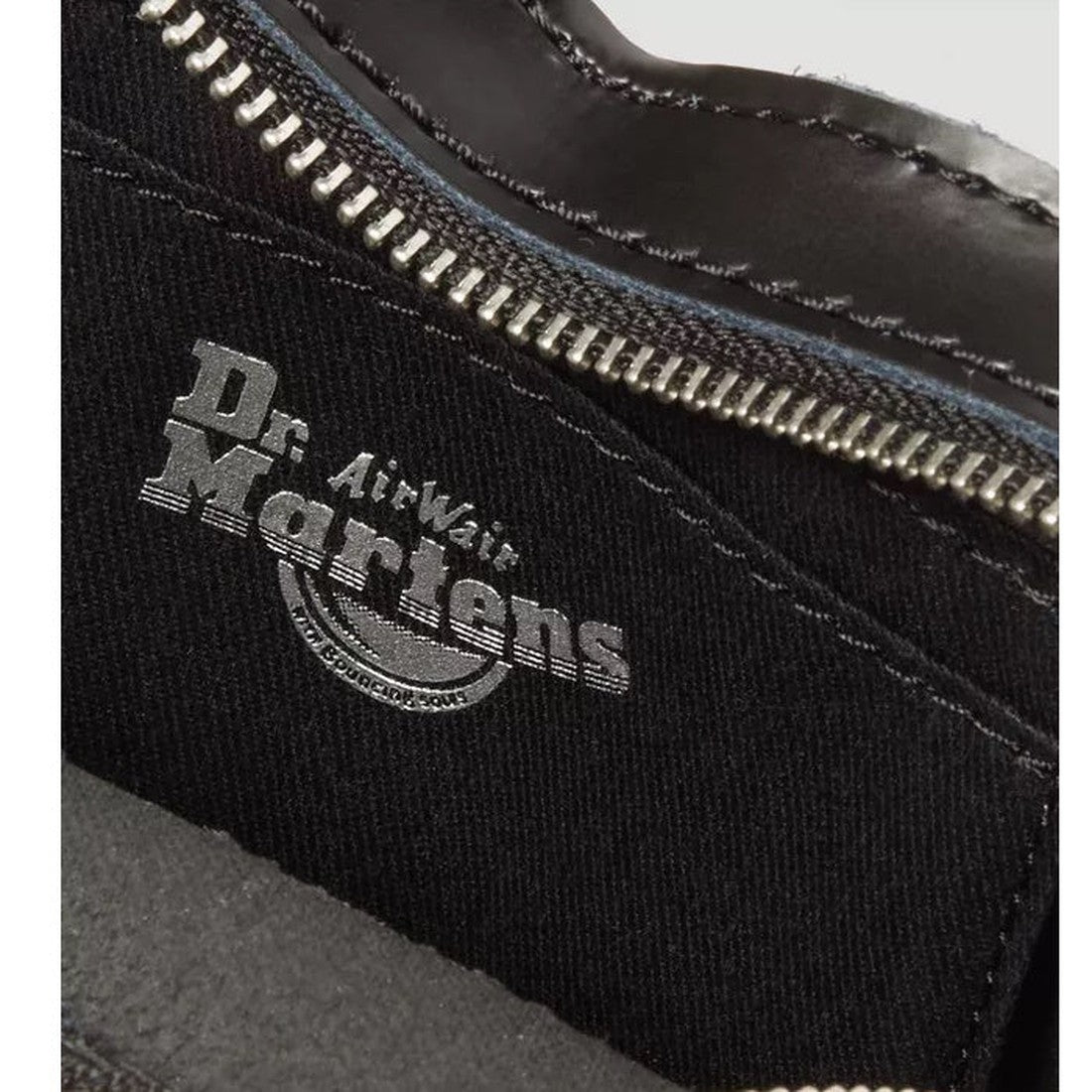 Close-up of a black Dr. Martens shoe with silver zipper and the “Dr. AirWair Martens” logo on the inner fabric, paired stylishly with a glossy Dr. Martens Mini Heart Leather Cross Body Black Patent Leather Satchel in the background., 190665640373