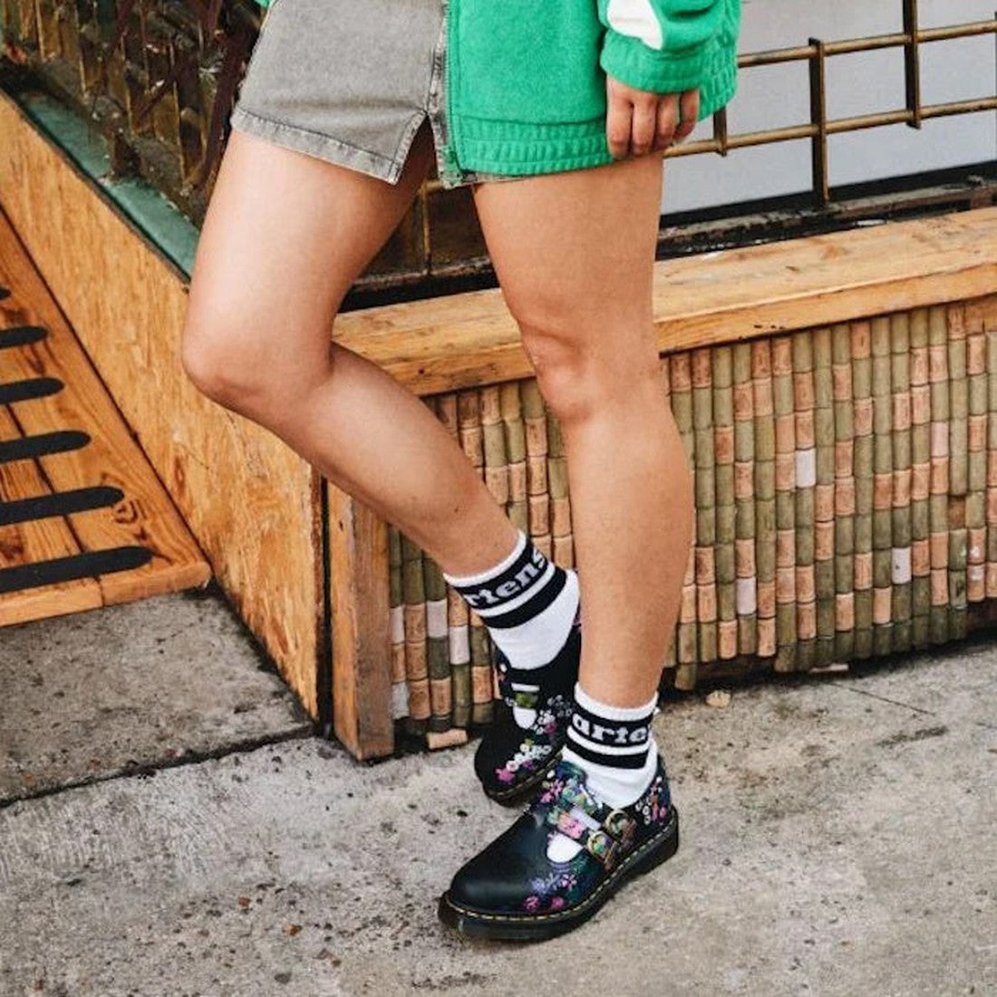 Only their legs are visible as a person stands outdoors in a green jacket, denim skirt, white socks with black lettering, and Dr Martens 8065 2 Strap Mary Jane Shoes in Vintage Floral Black., 190665573091, 190665573107, 190665573114, 190665573138, 1906655