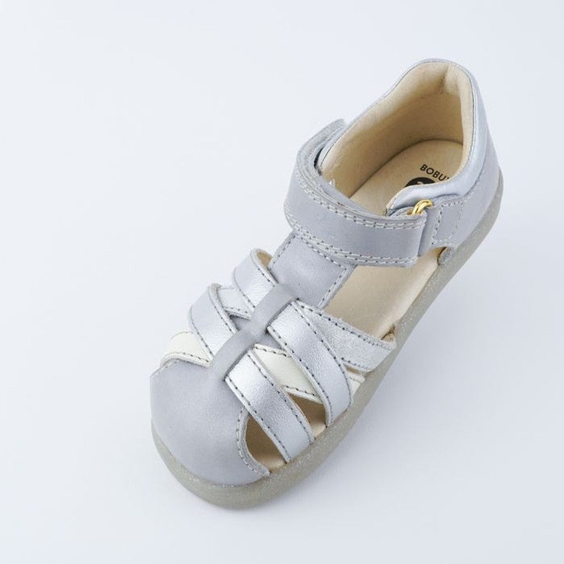 The Bobux iwalk Cross Jump Sandal Silver Pearl Closed Toe 636702 is a kids’ light gray leather sandal with crisscross straps, velcro ankle fastening, beige cushioned interior, and closed toe, shown on a plain white background., 9420062084045, 942006208405