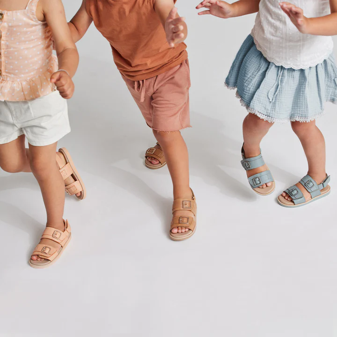 Three kids in summer outfits wear Pretty Brave Alex Leather Sandal Seaform Blue (sizes 29-33, clearance, not returnable) as they walk and play together on a white background, focusing on their legs and feet. Upper bodies mostly out of frame., 942103635768