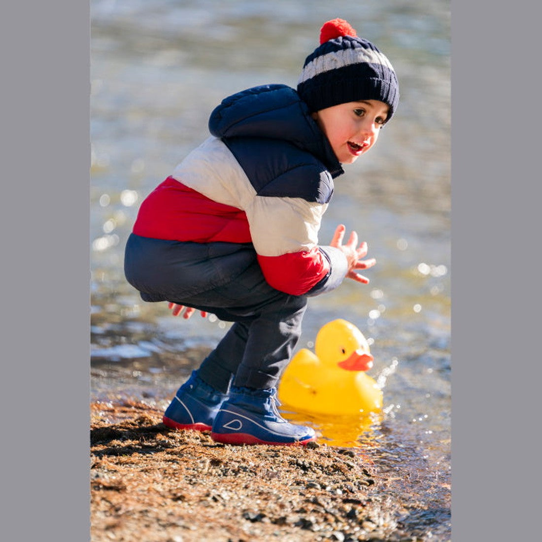 A young child in a striped winter jacket, hat, and blue boots squats by the water's edge, playing with a large yellow rubber duck—perfect for showcasing the Bobux Kid+ Splash Boot Waterproof Blueberry 839003., 9420068527911, 9420068527928, 9420068527935, 