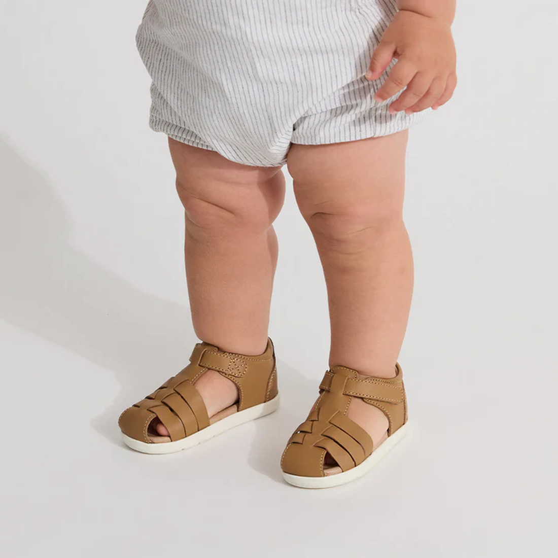A young child wearing light-colored shorts and Pretty Braver Billie First Walker Sandal Closed Back in tan stands on a white background, with only the legs and feet visible., 9420075334359, 9420075334342, 9421036357264