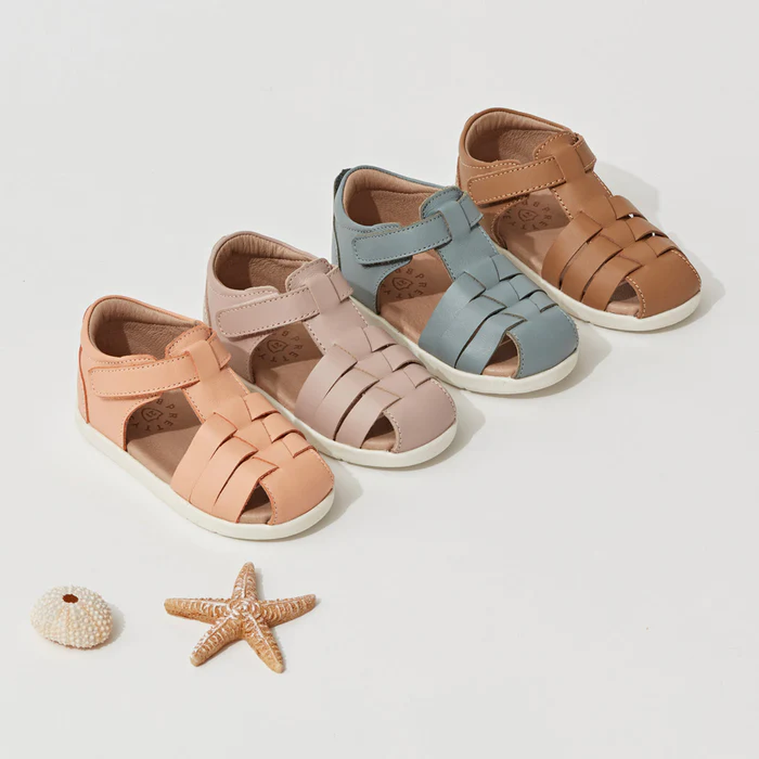 Four pairs of Pretty Braver Billie First Walker Sandal Closed Back Blush in peach, light brown, blue, and tan are lined up on a white background with a starfish and seashell. These lightweight, flexible sandals are ideal for little feet., 9420075334458, 9