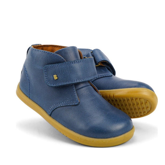 The Bobux Iwalk IW Desert Boot Midnight Blue 625217 is a pair of vintage-inspired children's boots in blue leather with tan rubber soles and a wide strap closure, displayed against a white background.