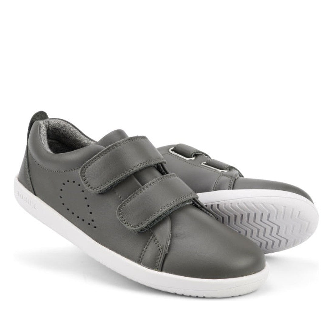 The Bobux Kids + Plus Grass Court Smoke Grey Velcro Trainers 832446 feature two gray velcro straps, white soles, side perforations, and a textured grip—ideal kids’ street shoes for everyday wear.