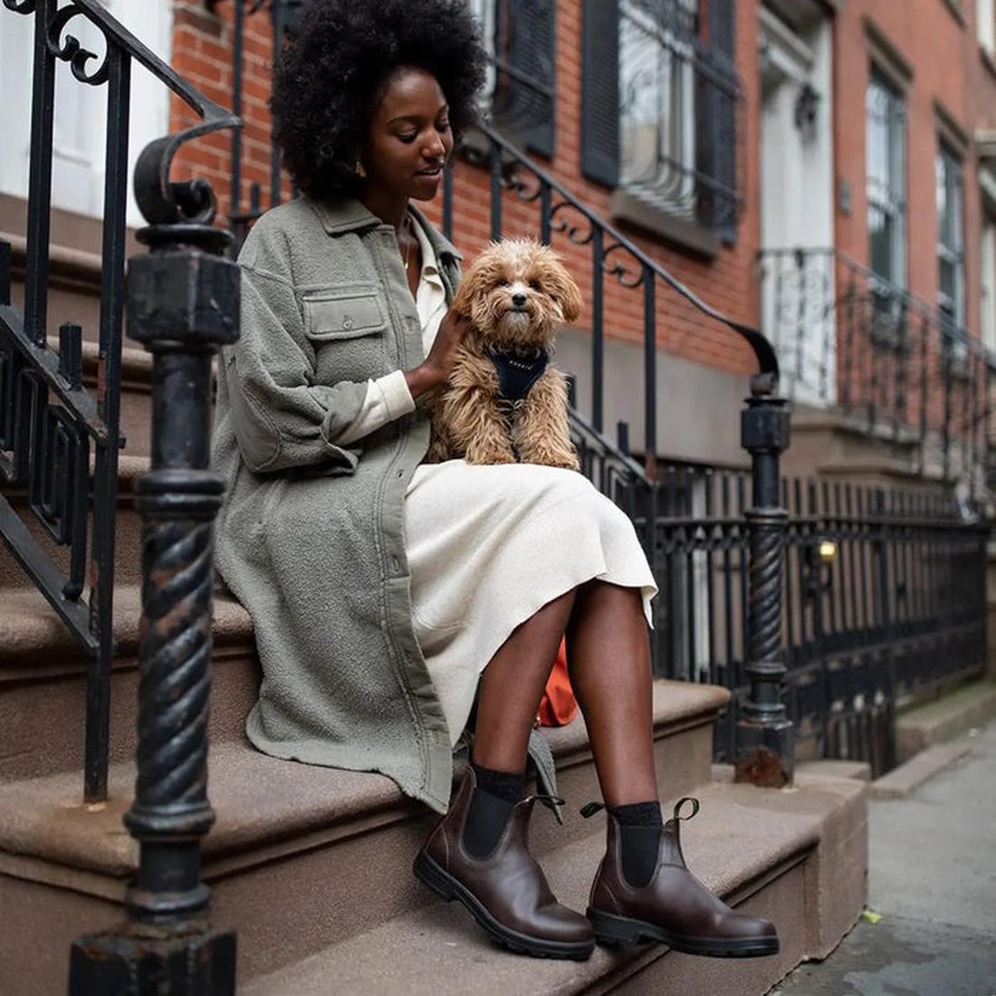 A cheerful woman in a long dress and green jacket pets her dog, wearing Blundstone 2116 VEGAN Brown Chelsea Boots with easy pull-on tabs.  ksnz, 9315891528689, 9315891528702, 9315891528726, 9315891528740, 9315891528764, 9315891528788, 9315891528801, 9315891528825