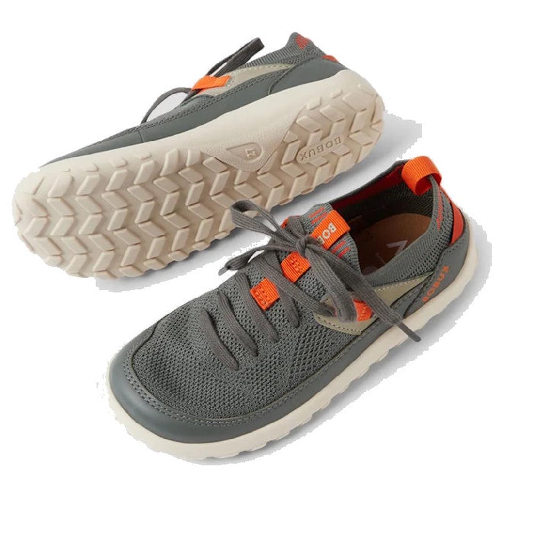 The Bobux Kid+ Meta Knit Barefoot Sneaker in Vetiver Green for juniors features a breathable knit upper, white textured sole, orange accents, and gray laces. One shoe lies on its side to display the sole; the other stands upright., 9420074836014, 94200748