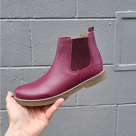 A hand holds the Clarks Kids Chelsea II Youths Leather Boot in maroon leather, featuring brown elastic side panels and a light-colored sole with Puddle Proof technology, shown against a gray brick wall.