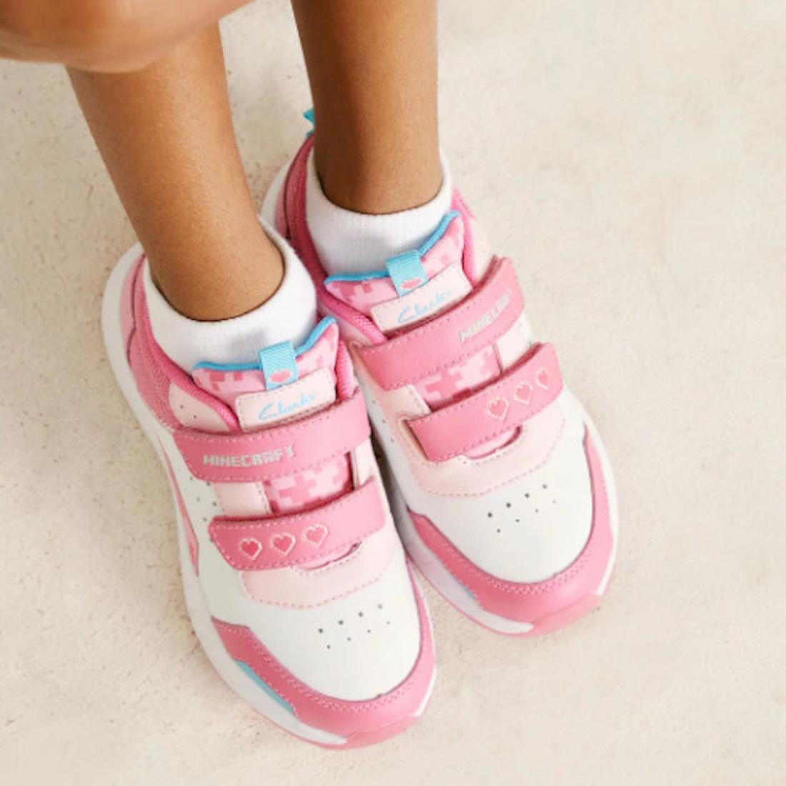 A child wears Clarks Hendy Pink/White Minecraft Trainer Shoes with Velcro straps, heart and pixel designs, and a comfy memory foam innersole, standing on a light-colored surface., 9336078918772, 9336078918789, 9336078918796, 9336078918802, 9336078918819, 