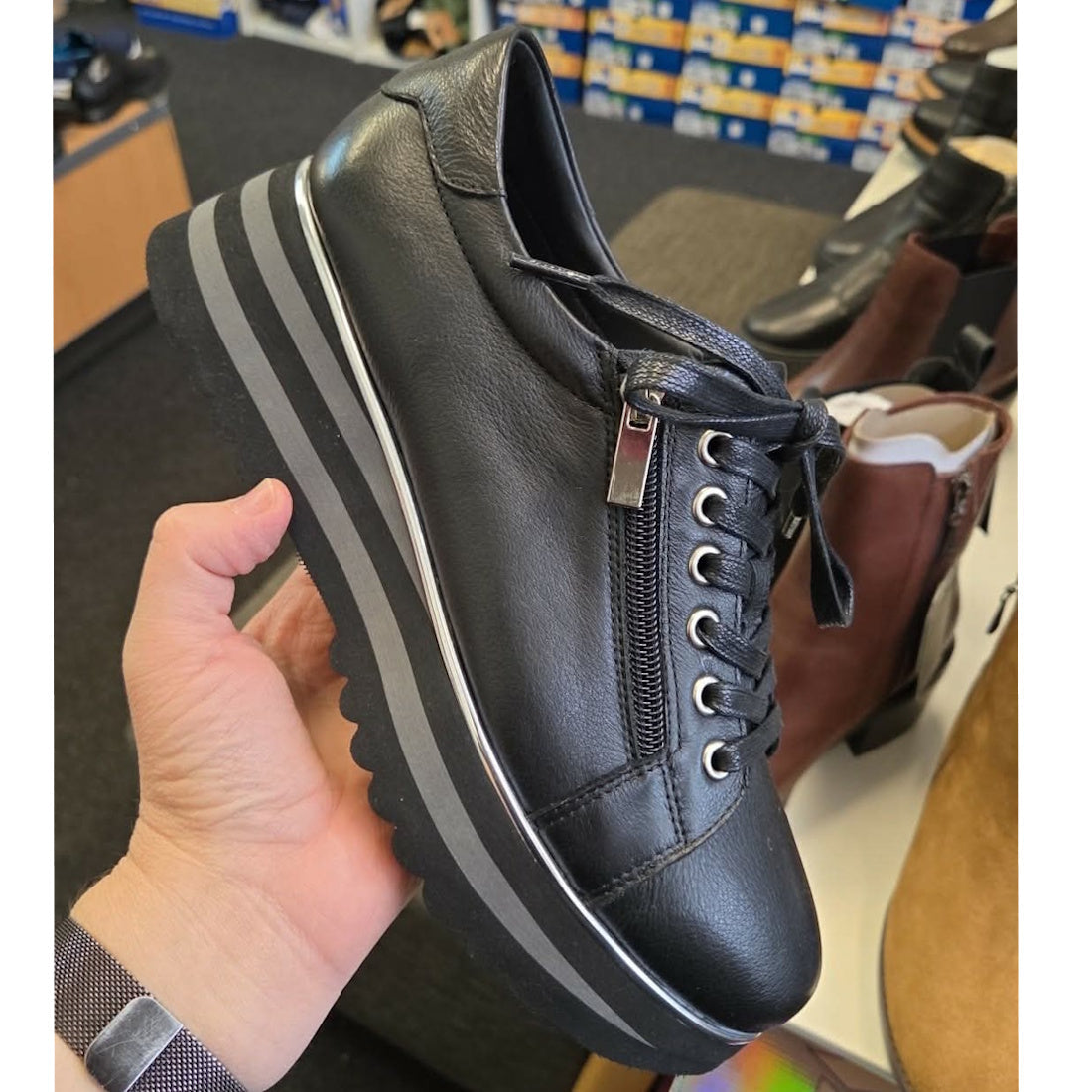 A hand holds the Django & Juliette Eammes Black Leather Platform Shoe Women's, featuring a thick striped sole, side zipper, and laces. Shoe shelves with various footwear boxes are visible in the background., 51089, 4434754993411, 4434756993428, 4434754993