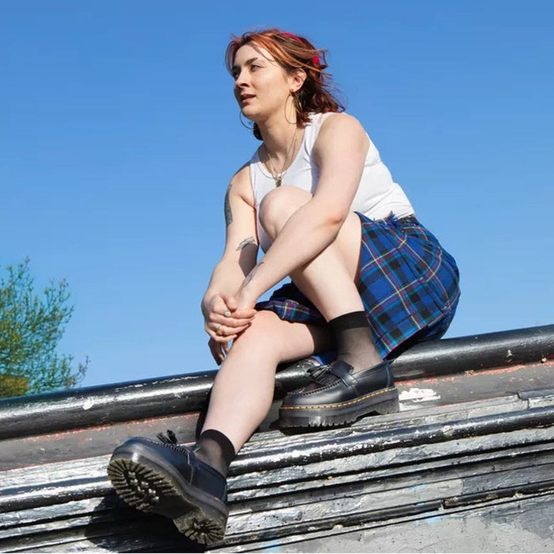 A person with light skin and red hair sits on a ledge outside under a clear blue sky, wearing a white tank top, blue plaid skirt, black socks, and Dr Martens Adrian Quad Black Smooth Loafer Shoes., 30429, 30437, 190665522402, 190665522419, 190665522426, 1