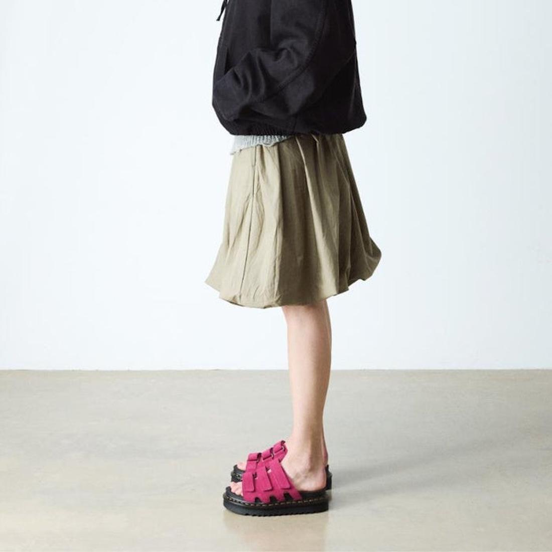 A person stands indoors wearing a black jacket, khaki skirt, and Dr Martens Raine Fuchsia Pink Greasy Suede Leather Slides with thick EVA Zebrilus soles. The background features a plain light wall and beige floor; face not shown., 47985, 190665750270, 190