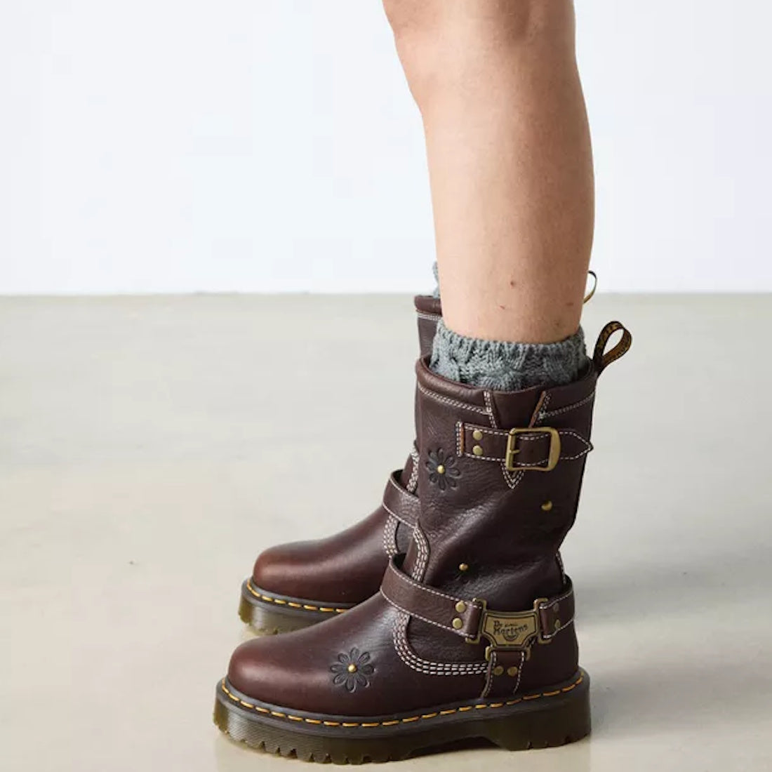 Someone wearing gray knit socks and Dr Martens Anistone Hi Flower Bex Grizzly Dark Brown boots, featuring buckles, decorative stitching, and floral details, stands on a light floor with a plain background., 52349, 190665773651, 190665773668, 190665773682,