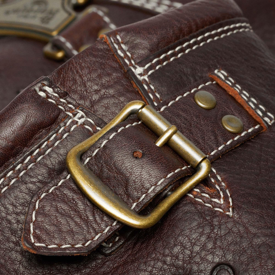 Close-up of the Dr Martens Anistone Hi Flower Bex Grizzly Dark Brown bag, showing textured brown leather, white stitching, a brass buckle, and metal rivets. Stitching and hardware details are clearly visible., 52349, 190665773651, 190665773668, 1906657736