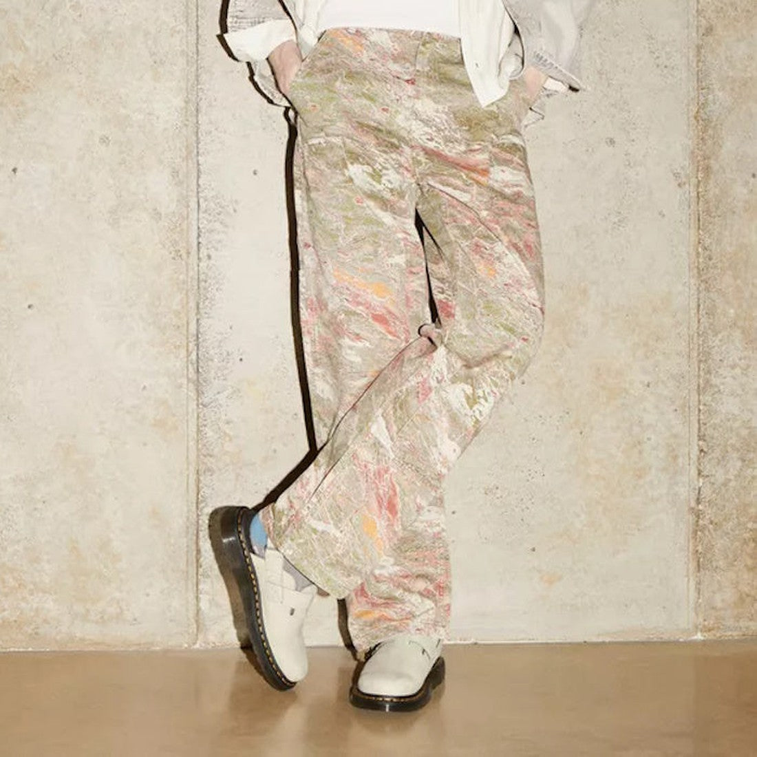 A person stands against a beige wall with legs crossed, wearing loose marbled pants in pink, green, and beige tones, a light-colored jacket, and Dr Martens Jorge II Warm Stone Buttersoft Sling Back Sandal Covered Toe. The photo is cropped above the should