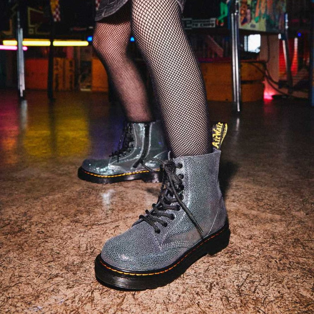 A person in fishnet tights wears Dr Martens Kids 1460 J Iridescent Metallic Kidray Boots with black soles, standing on a textured floor under dim indoor lighting with colorful lights glowing in the background., 190665432046, 190665432060, 190665432084, 16