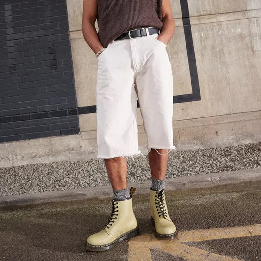 A person stands against a concrete and brick wall, hands in pockets, wearing a brown sleeveless shirt, white frayed knee-length shorts, gray socks, and yellow Dr. Martens 1460 Smooth Pale Olive 8 up Leather Boots., 190665531824, 190665531831, 190665531848