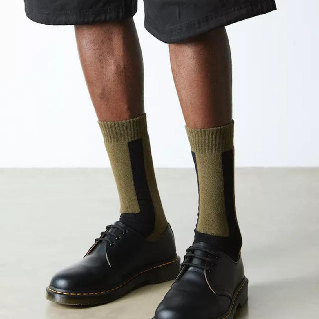 A person in black shorts, cozy Dr Martens Socks Double Doc Sock Olive Unisex, and classic black leather lace-up shoes with sturdy soles.  ksnz, 190665728279, 190665728262