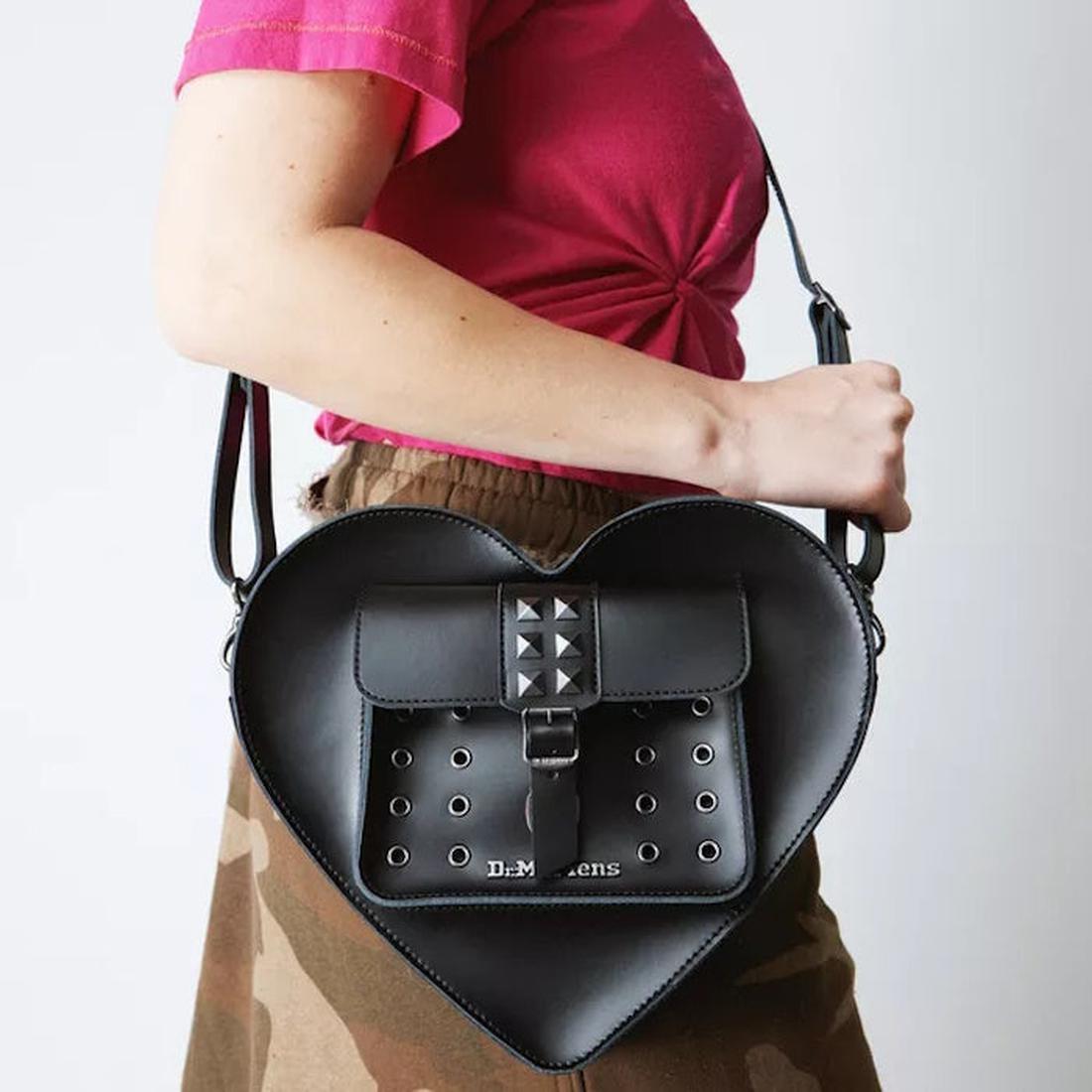 A young person with a pink top and camo skirt holds the Dr Martens Heart Shaped Bag, shown with buckle closure and silver studs.  ksnz, 190665728408