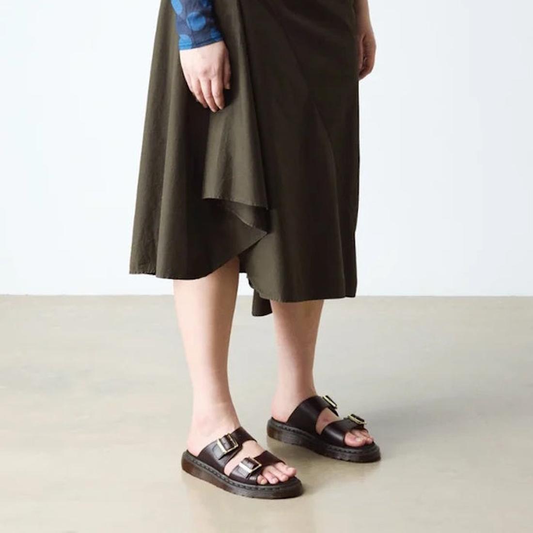 A person wears a dark olive asymmetrical midi skirt and black Dr Martens Josef Charro Atlas Slide Sandal Leather on a light floor, with only the lower half of their body visible., 52357, 190665783841, 190665783858, 190665783872, 190665783889, 190665783896