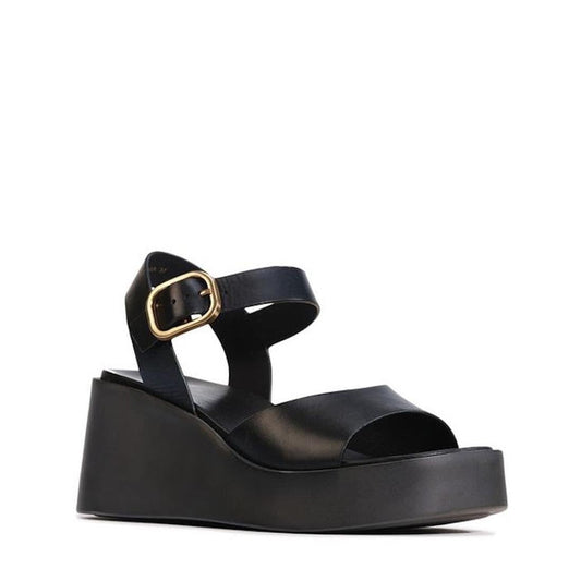 The EOS Ariya Black Leather Dressy Wedge Platform Sandal features a thick sole, open toe, ankle strap with gold buckle, and hand-burnished leather for a minimalist yet modern look that blends comfort and style.