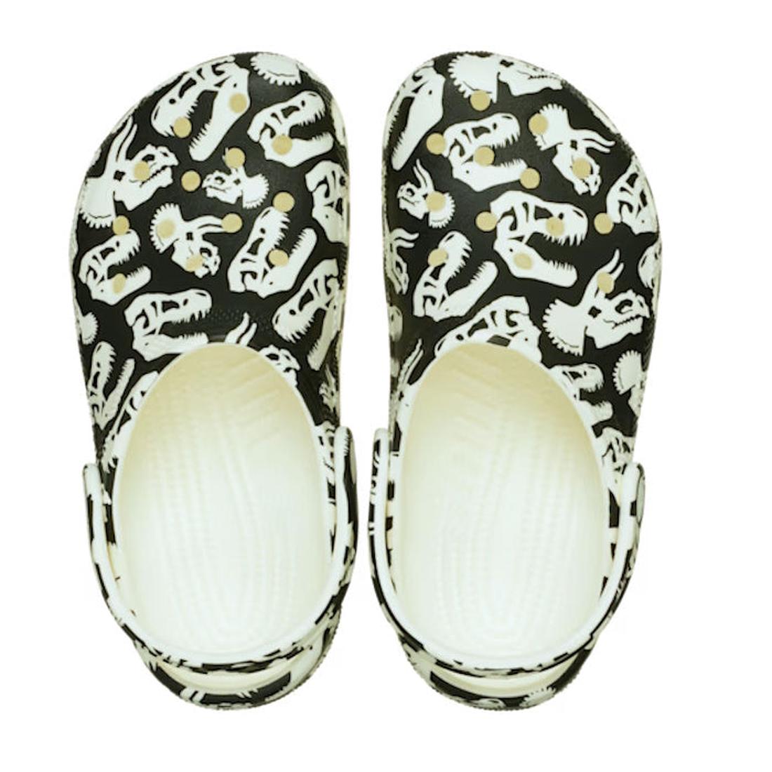 The Crocs Kids - Youths Classic Glow Dino Clog K Black Sandal features black clogs with white dinosaur skull patterns, glow in the dark details, and adjustable heel straps, shown from above on a white background., 198445261459, 198445261466, 198445261473,