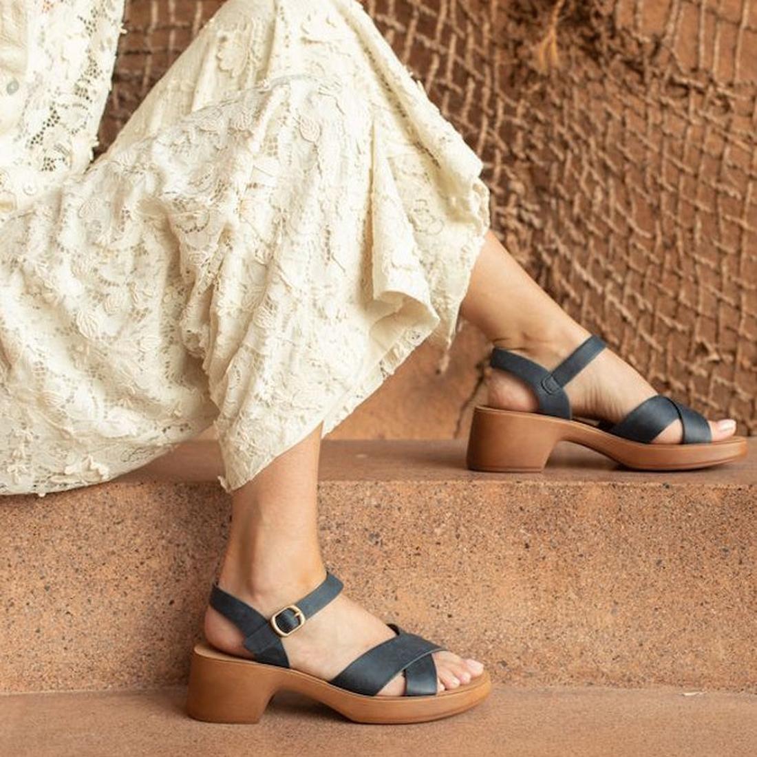 A woman in a cream lace dress sits outdoors, wearing Los Cabos Alegra Navy Wedge Sandals—secure backstrap and comfy platform sole.  ksnz