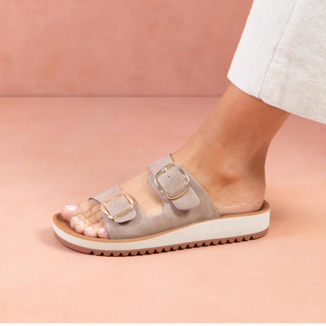 A person in light pants wears the Los Cabos Naara Taupe Adjustable Womens Summer Slide Sandal, featuring two gold buckles, on a pink surface. The side view highlights the sandal’s vegan leather design., 9321191159003, 9321191159010, 9321191160078, 9321191