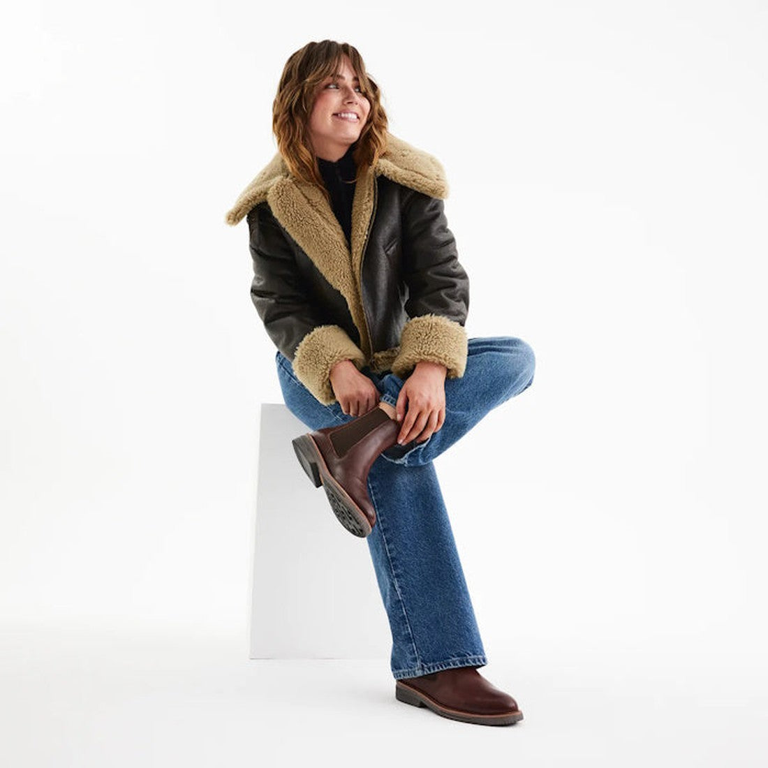 A person wearing a shearling-lined black leather jacket, blue jeans, and Rollie Womens Chelsea Rise Vintage Mocha Leather Boots sits on a white block against a plain white background, smiling with one leg crossed over the other., 9339554341682, 9339554341