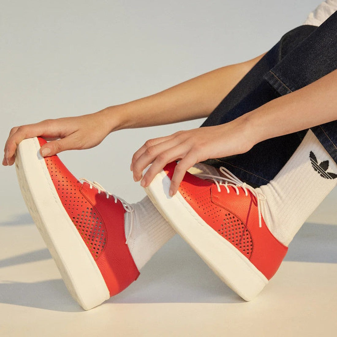 A person in dark blue jeans and white socks with a black Adidas logo sits on the floor, slightly bent forward, touching their Rollie Womens Derby City Punch Red Leather Trainers with white soles., 9781760775599, 9781760775605, 9781760775612, 9781760775629