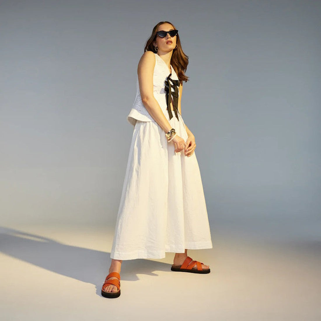 A woman in a sleeveless white top with black bow detail, long white skirt, sunglasses, bracelets, and Rollie Womens Rhodes Cross Slide Tangerine Leather sandals poses confidently in a minimalist studio with soft lighting., 9781760767389, 9781760767396, 97