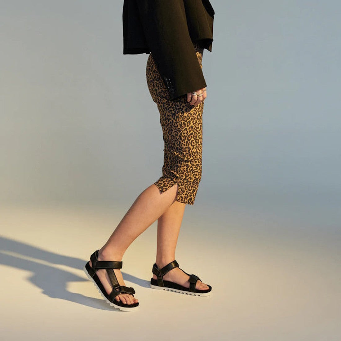 A person stands on a neutral background, facing left. Only their lower body is visible, showcasing Rollie Womens Sandal Tooth Wedge Black with adjustable sheep leather straps, animal print capri pants, and a black jacket., 9339554310381, 9339554310398, 93