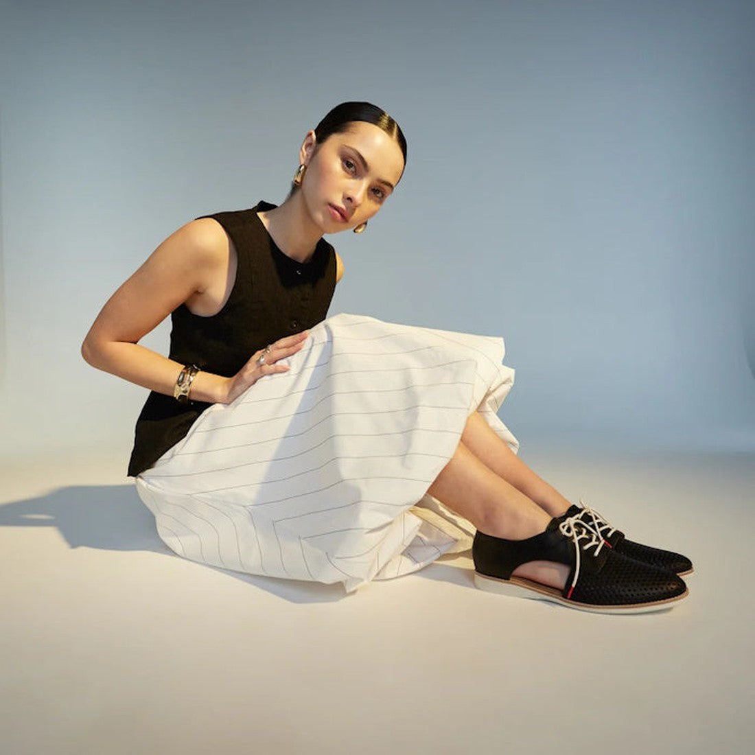 A woman with dark hair in a bun sits on the floor against a plain background, wearing a black sleeveless top, white striped skirt, and Rollie Womens Sidecut Punch Black Leather Shoes. She looks towards the camera with a neutral expression., 9339554249179,