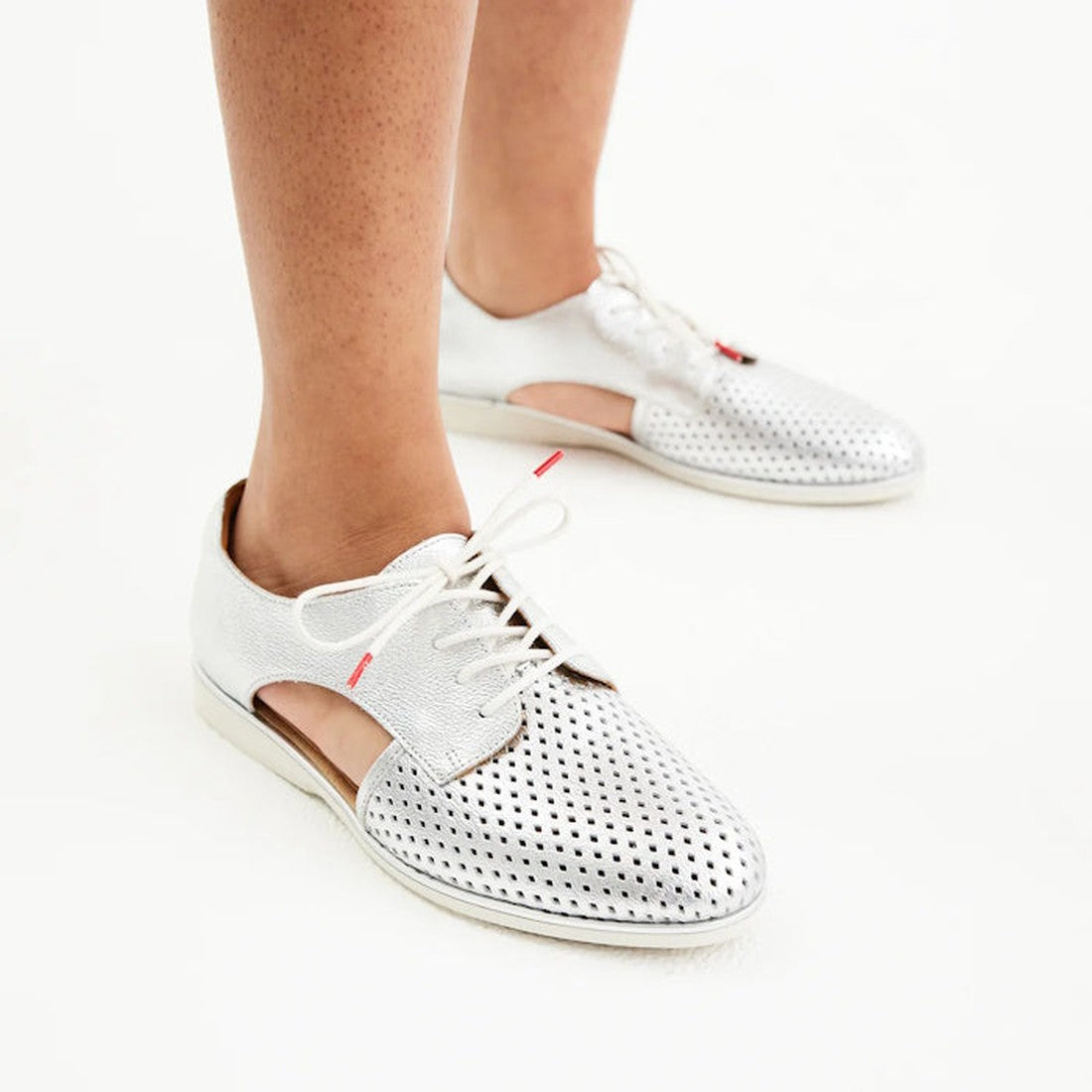A person wears the Rollie Womens Sidecut Punch Silver Metallic Leather Shoe on a white surface. This breathable shoe features cut-out sides, white laces, a low heel, and an anti-slip rubber outsole for comfort and stability., 9339554231341, 9339554231358,