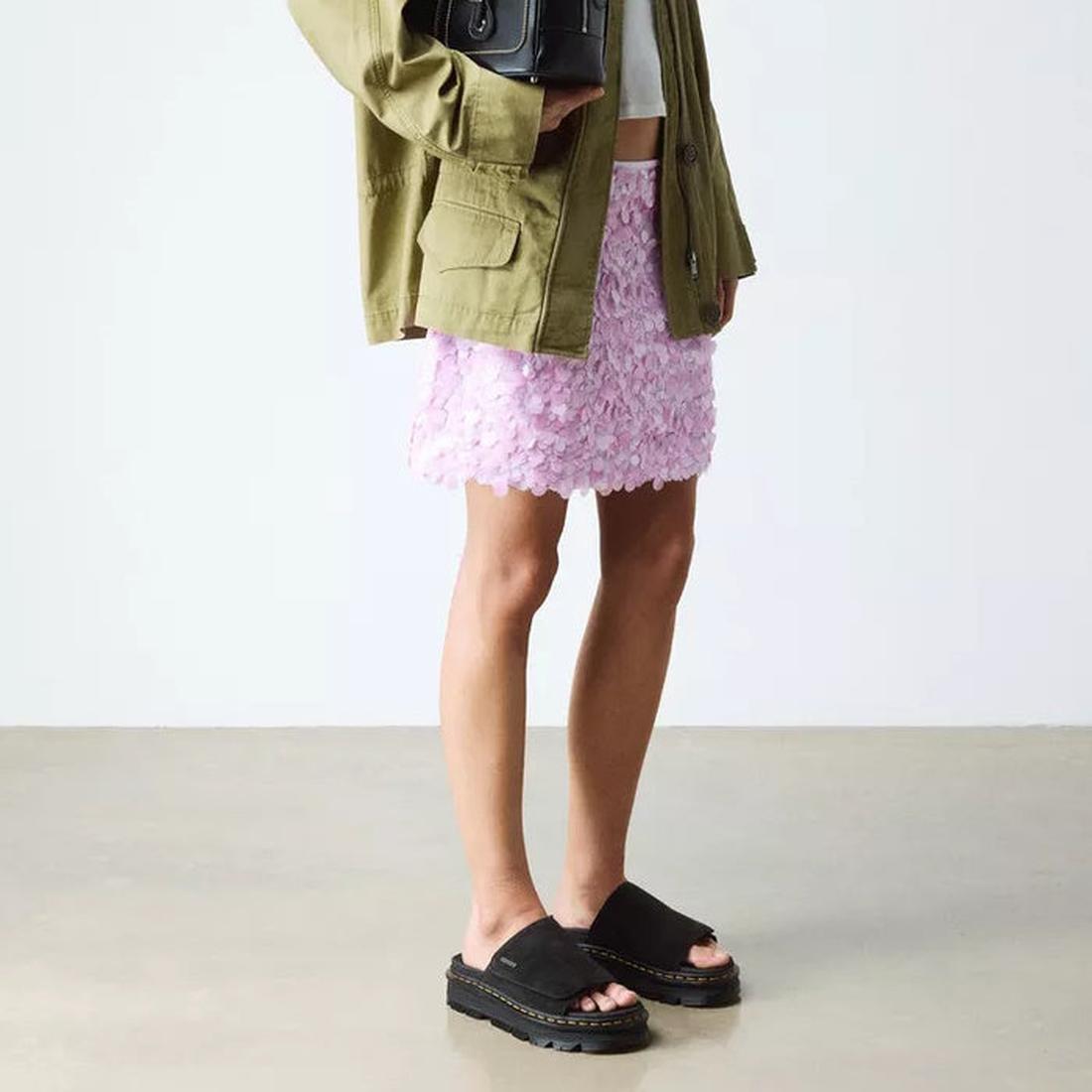 Person wearing an olive green jacket, pale pink textured mini skirt, and Dr Martens Zebzag Black EH Suede Slide Sandals with air-cushioned sole, standing indoors on a light floor against a white background., 190665790689, 190665790696, 190665790702, 19066