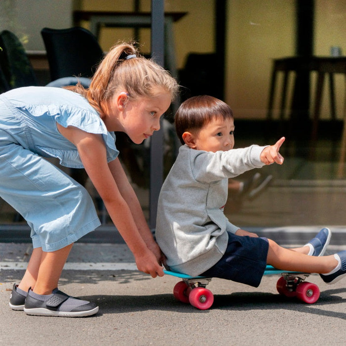 A young girl in a light blue outfit, wearing Bobux Iwalk Lo Dimension III Blue Navy Trainer 638506 Unisex shoes, pushes a boy on a skateboard with pink wheels. The boy, in a gray sweater and shorts, points ahead as they play outdoors near glass., 94200620
