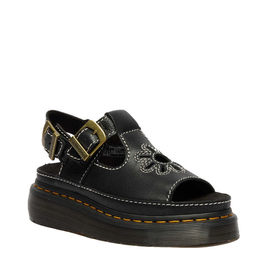 The Dr Martens Dunnet Flower Sandal Black Quad Platform features a black full-grain leather upper with floral cutouts, peep toe, white stitching, adjustable ankle strap with gold buckle, and a thick black sole accented by yellow stitching.