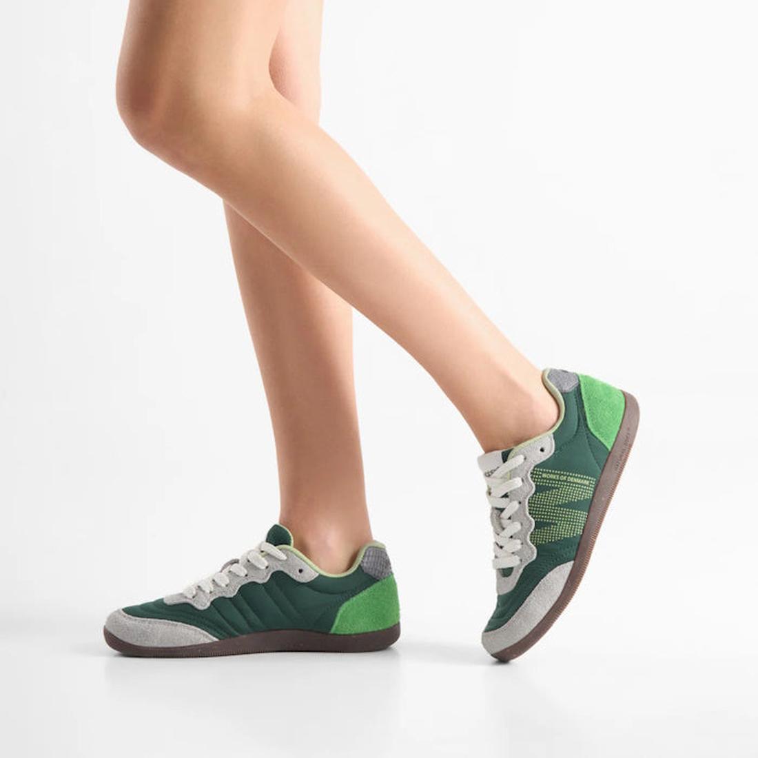 A person wears Woden Nanna Apple Green Multi Lace Up Sneakers, featuring green and gray accents with white laces, standing on one leg with the other bent slightly in front against a plain white background., 50250, 5713326697834, 5713326697841, 57133266978