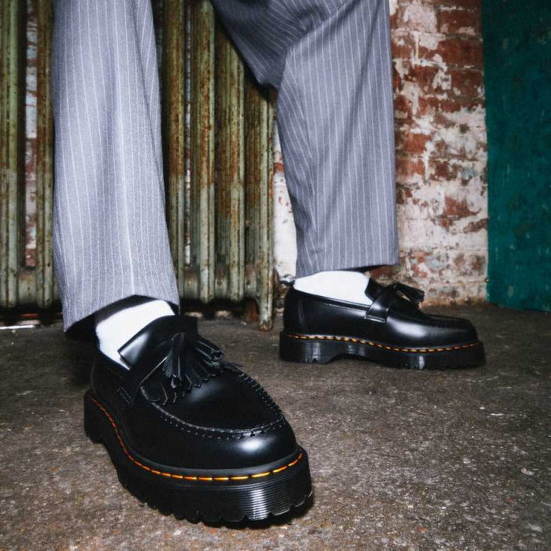 Someone stands before a distressed brick wall and old radiator, wearing Dr Martens Adrian Bex Black Smooth Platform Loafer Shoes (CLEARANCE), paired with white socks and gray pinstripe trousers., 183681, 190665420241, 190665420166, 190665420173, 190665420
