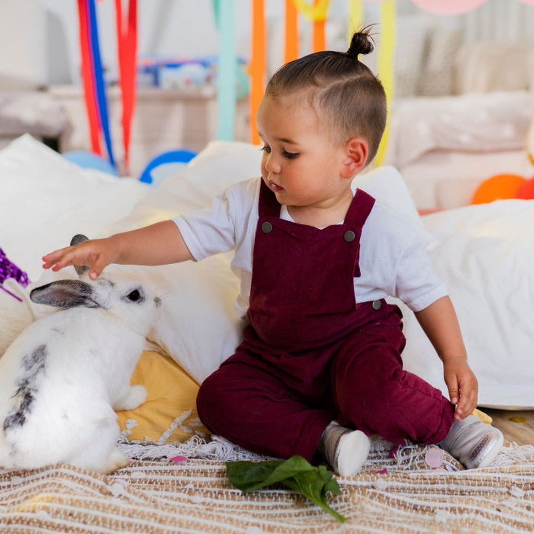 A toddler in maroon overalls, wearing Bobux Explorer Go Rose Gold First Walker Crawler 501015B shoes, gently pets a white rabbit with black spots on a yellow blanket with leafy greens, while colorful streamers hang in the background., 9420075704473, 94200
