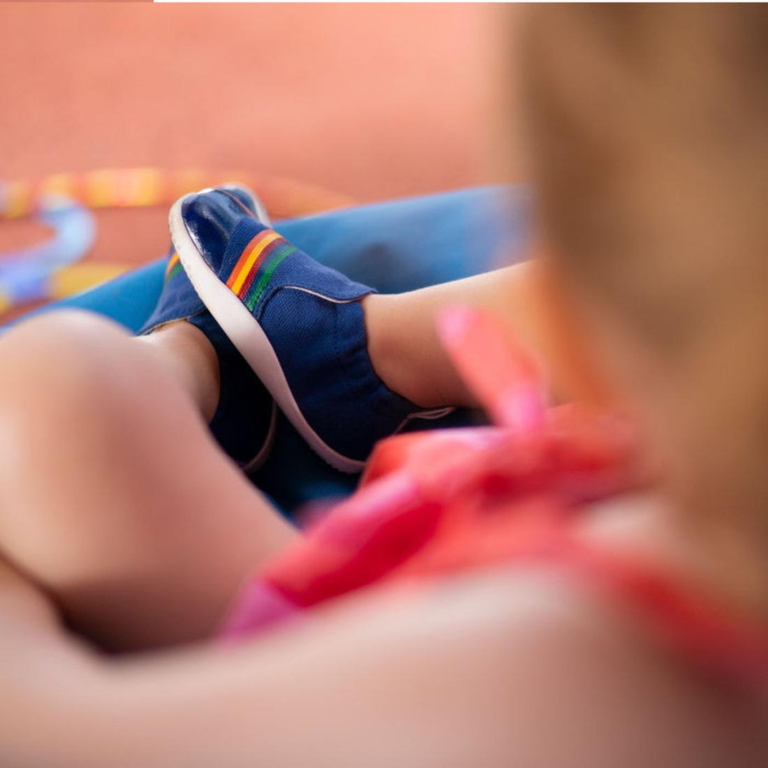 A young child wears Bobux Xplorer Scamp Crawler Blueberry Red Rainbow 501711 canvas shoes, relaxing with crossed legs. The bright outfit and the colorful, flexible sole of these first walking shoes stand out in the scene., 9420068534483, 9420068534490, 94