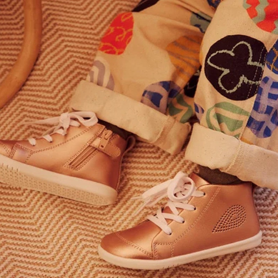 A child in IWalk IW Bobux Alley Oop Boot Leather Toddlers Rose Gold Metallic 637113 sits on a beige textured carpet, wearing white-laced boots and beige pants with colorful abstract designs., 9420071321681, 9420071321698, 9420071321704, 9420071321711, 942