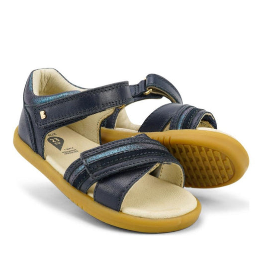 The Bobux iwalk IW Magic Girls Leather Sandal in navy features open toes, adjustable Velcro straps, beige cushioned insoles, and flexible light brown rubber soles. One sandal stands upright while the other is laid on its side.