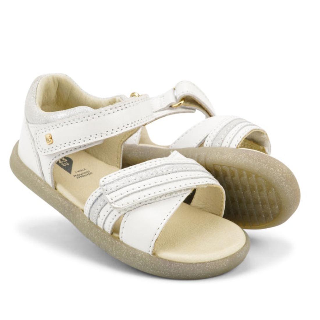 The Bobux iwalk IW Magic Girls Leather Sparkle Sandal in white features velcro straps and beige soles, with one sandal upright and the other leaning against it, showcased on a white background.