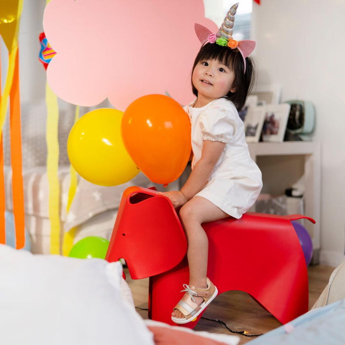 A young child in white sits on a red elephant toy, holding yellow and orange balloons, and wears Bobux IW Iwalk Mirror Pale Gold Girls Open Toed Sandals. The room is filled with colorful balloons and a large pink cloud cutout., 9420068524088, 942006852409