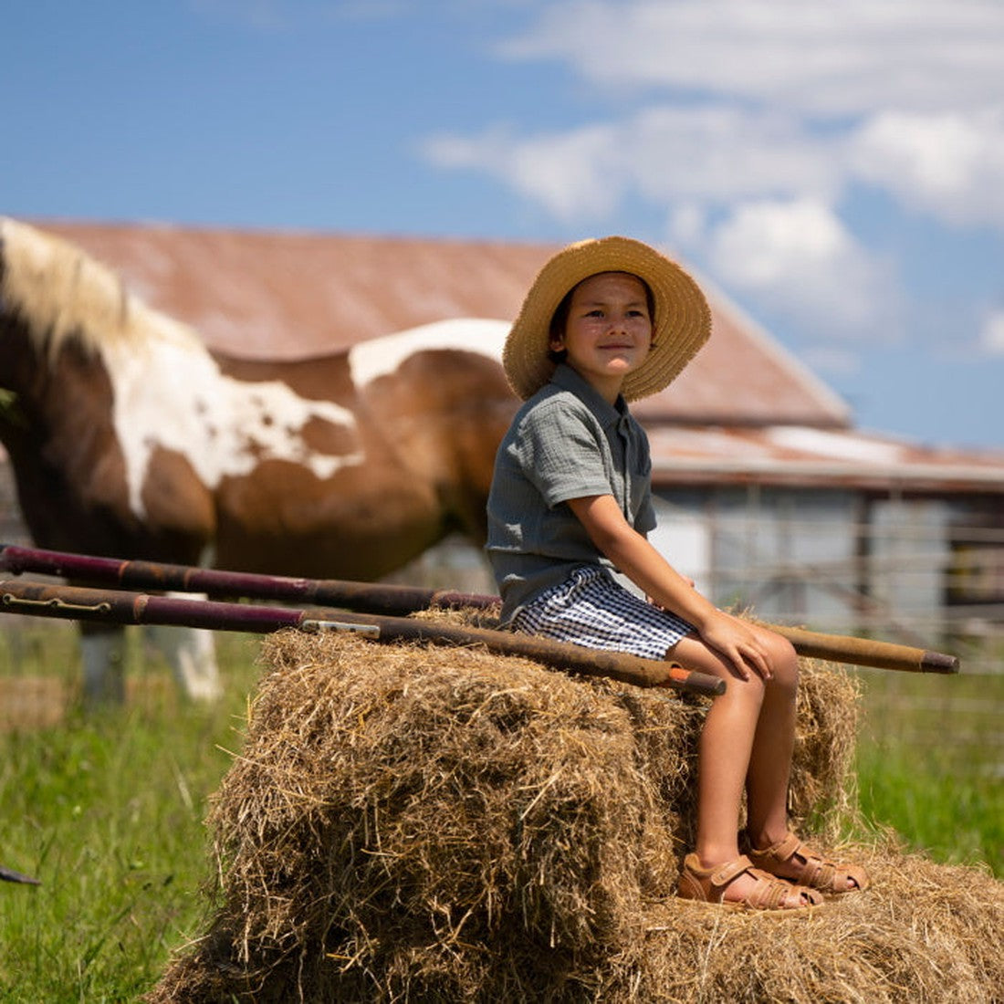 A young child in a straw hat and Bobux Kid+ Plus Roam Caramel Closed Toe Sandal (830506A) sits on a hay bale on a sunny farm, with a brown and white horse and rustic barn in the background., 9420062049952, 9420062049969, 9420062049976, 9420062049983, 9420