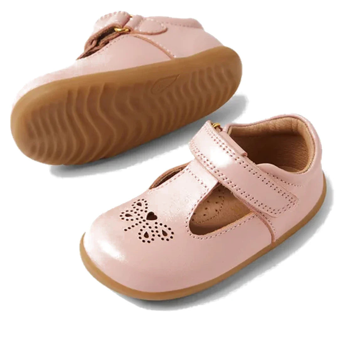 The Bobux Step Up T-Bar Rosie First Walker Shoe in Ballet Pink features pink leather, tan rubber soles, a T-strap, and heart and flower cutouts on the toe. One Mary Jane flat is upright while the other displays its sole., 51161, 9420074849946, 94200748499