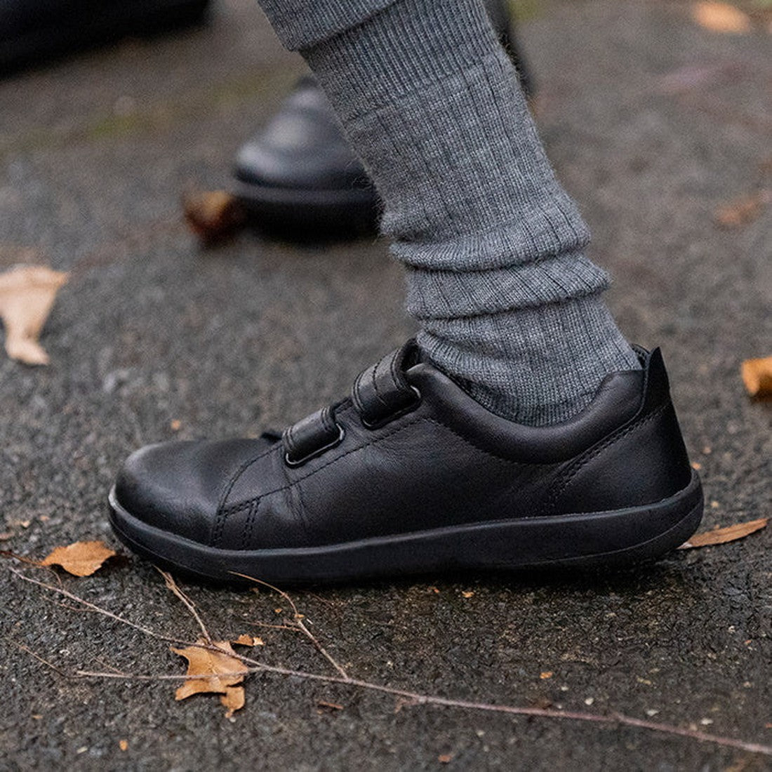 A close-up of a child’s foot in the Bobux Kid+ Black School Leather Shoe Venture Trainer with two Velcro straps, gray socks, and a non-marking sole, standing on pavement scattered with dry leaves., 9420068510289, 9420068510296, 9420068510302, 942006851031