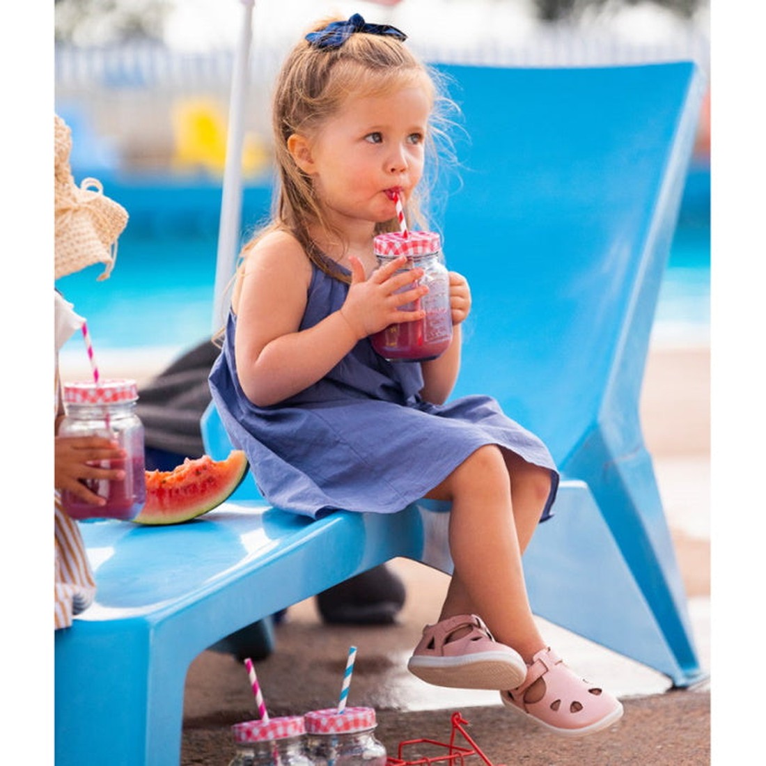 A young girl in a blue dress sits by the pool on a blue chair, sipping from a mason jar with a red-striped straw. She wears Bobux IWalk IW Zap II Sandal Pink Shimmer sandals, has a navy bow, and a slice of watermelon beside her., 9420068509245, 9420068509